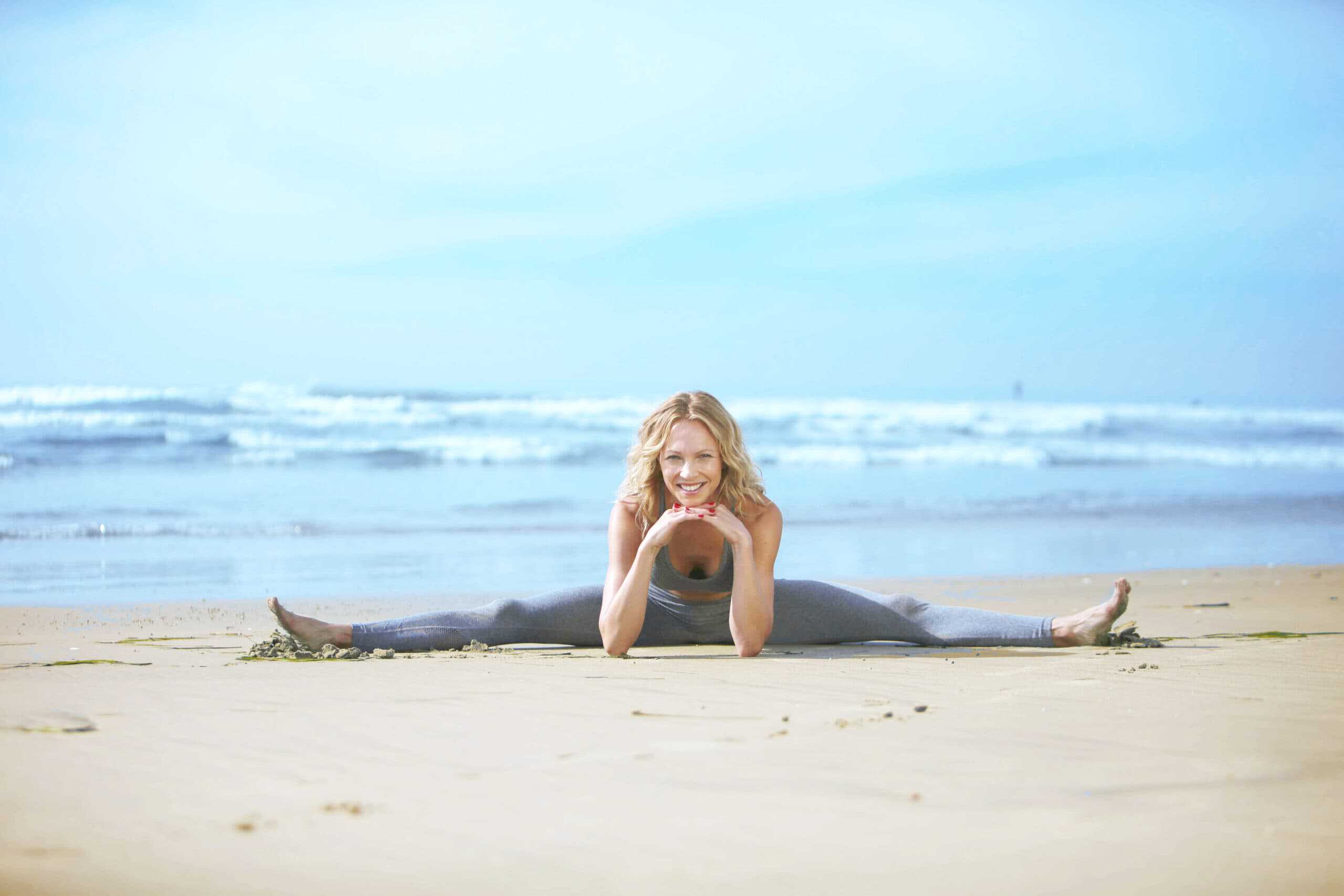 Auf dem Bild sieht man Birte Glang, wie sie glücklich am Strand im Spagat sitzt. Diese Pose zeigt eine beeindruckende Flexibilität und ist ein gutes Beispiel für fit bleiben in der Schwangerschaft. Birte ist somit bereit für Sport nach der Rückbildung.